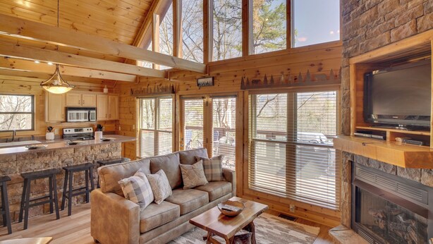 Un intérieur de chalet chaleureux avec une cheminée en pierre, un coin repas en bois et de grandes fenêtres offrant une vue sur l’extérieur.