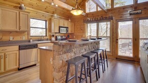 Une cuisine rustique aménagée dans une cabane en bois, avec un îlot central en pierre, des tabourets de bar et une grande fenêtre.