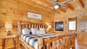 Une chambre dans une cabane en bois, avec un lit, deux tables de chevet, un ventilateur de plafond et un téléviseur fixé au mur.