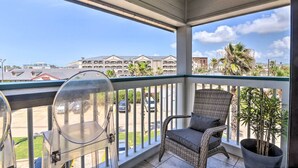 A balcony with a wicker chair and a potted plant, overlooking a parking lot and buildings. - Steps to Sand + 2 Pools! Galveston Coastal Getaway (Galveston)