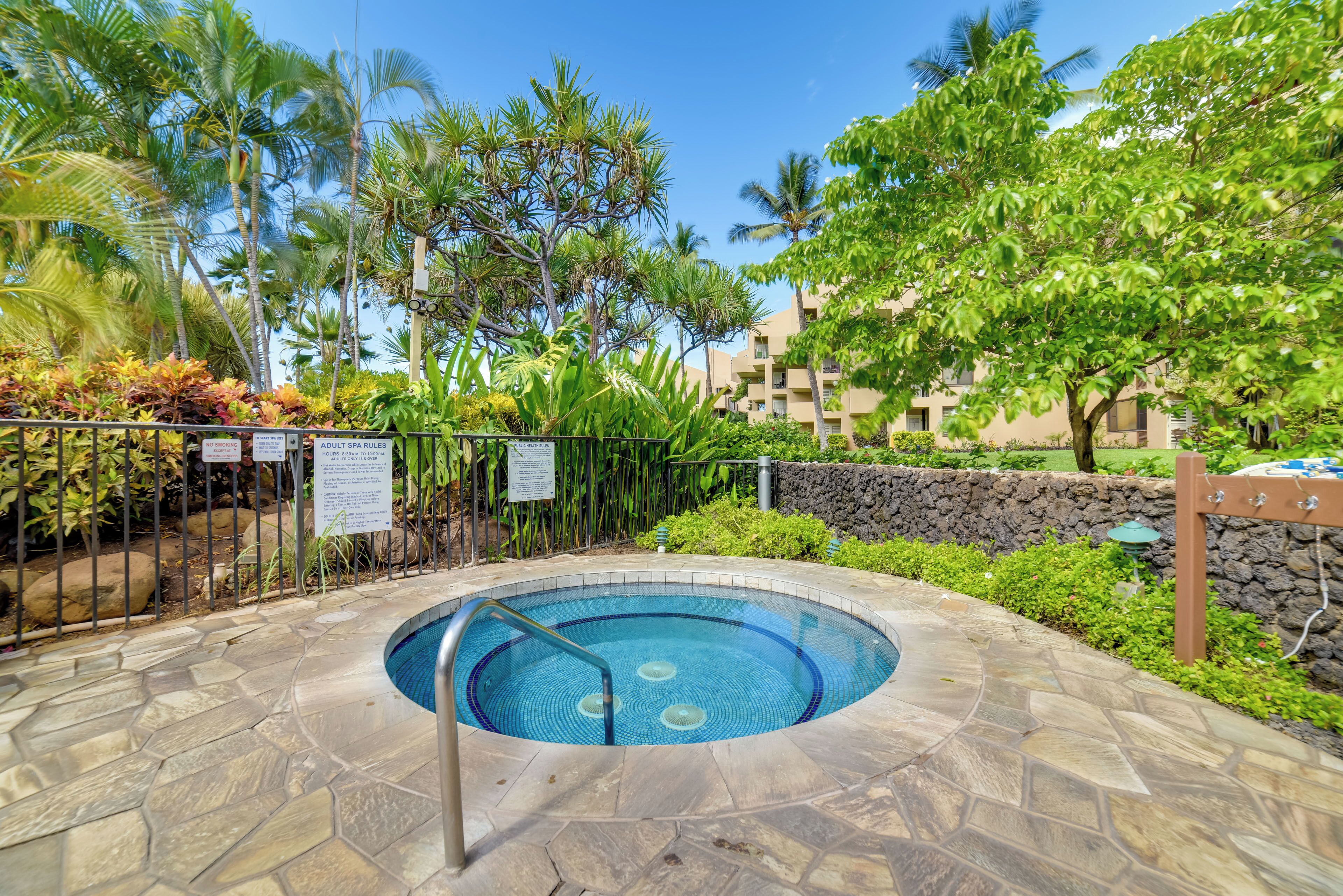 A circular hot tub surrounded by a stone patio, with a pool in the background, and a fence with signs indicating spa rules.