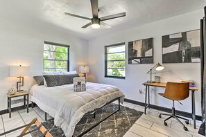 Ein Schlafzimmer mit einem Bett, zwei Nachttischen, einem Schreibtisch, einem Stuhl, einem Deckenventilator und zwei Fenstern mit Blick auf Grünflächen.