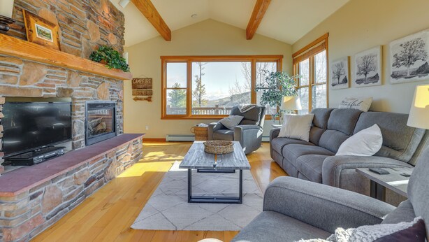 A living room with a stone fireplace, a flat-screen TV, and a large window offering a view of the outdoors. - Private Hot Tub: Scenic Granby Vacation Rental! (Granby)