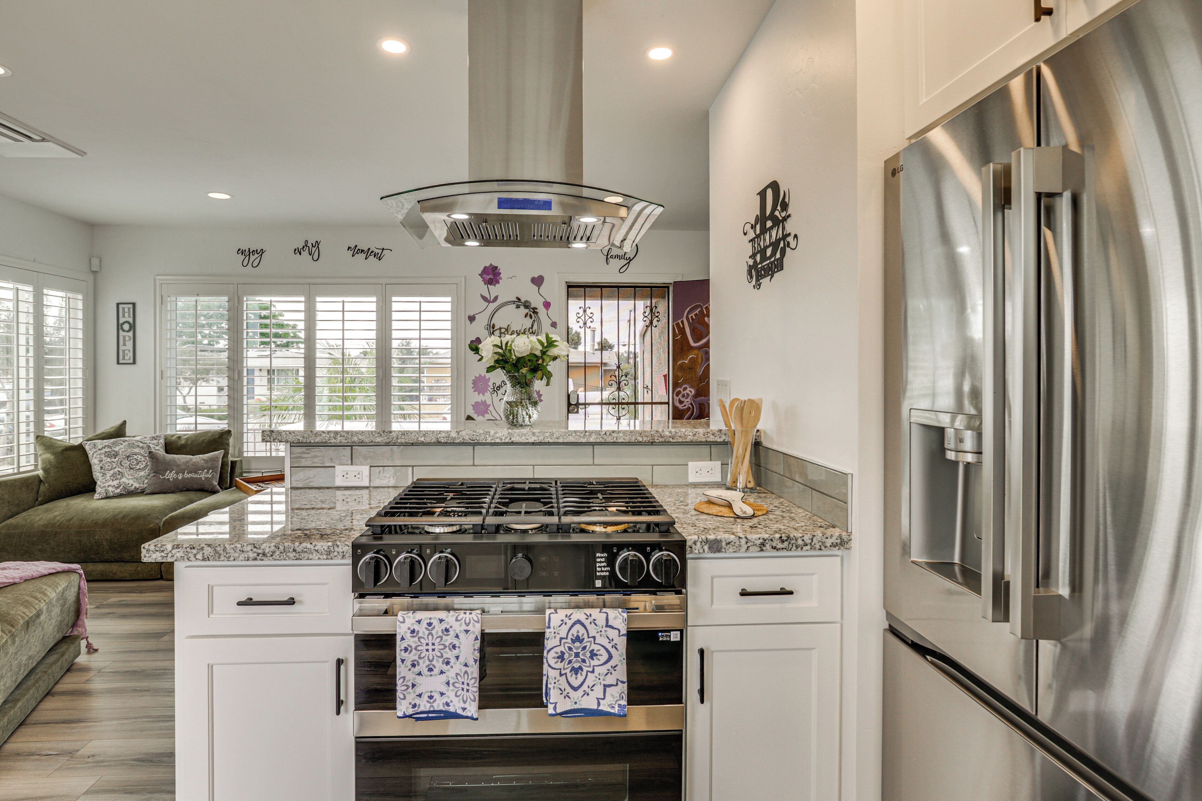 A modern kitchen with a stainless steel refrigerator, a gas stove, and a built-in oven. There is a large window with blinds, a decorative wall with text and flowers, and a green sofa in the living area.