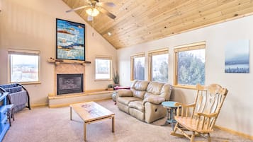 A cozy living room with a fireplace, a ceiling fan, and large windows.