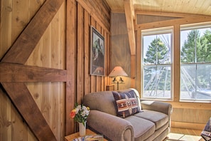 A cozy room with wooden walls, a sofa with patterned cushions, a lamp, and a window with a view of trees.