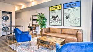 En moderne stue med en brun lædersofa, blå stole, et træbord og et tæppe. I baggrunden ses en vaskemaskine, en stige og en potteplante. To indrammede plakater på væggen viser ordene 'HAPPINESS' og 'PATIENCE'.