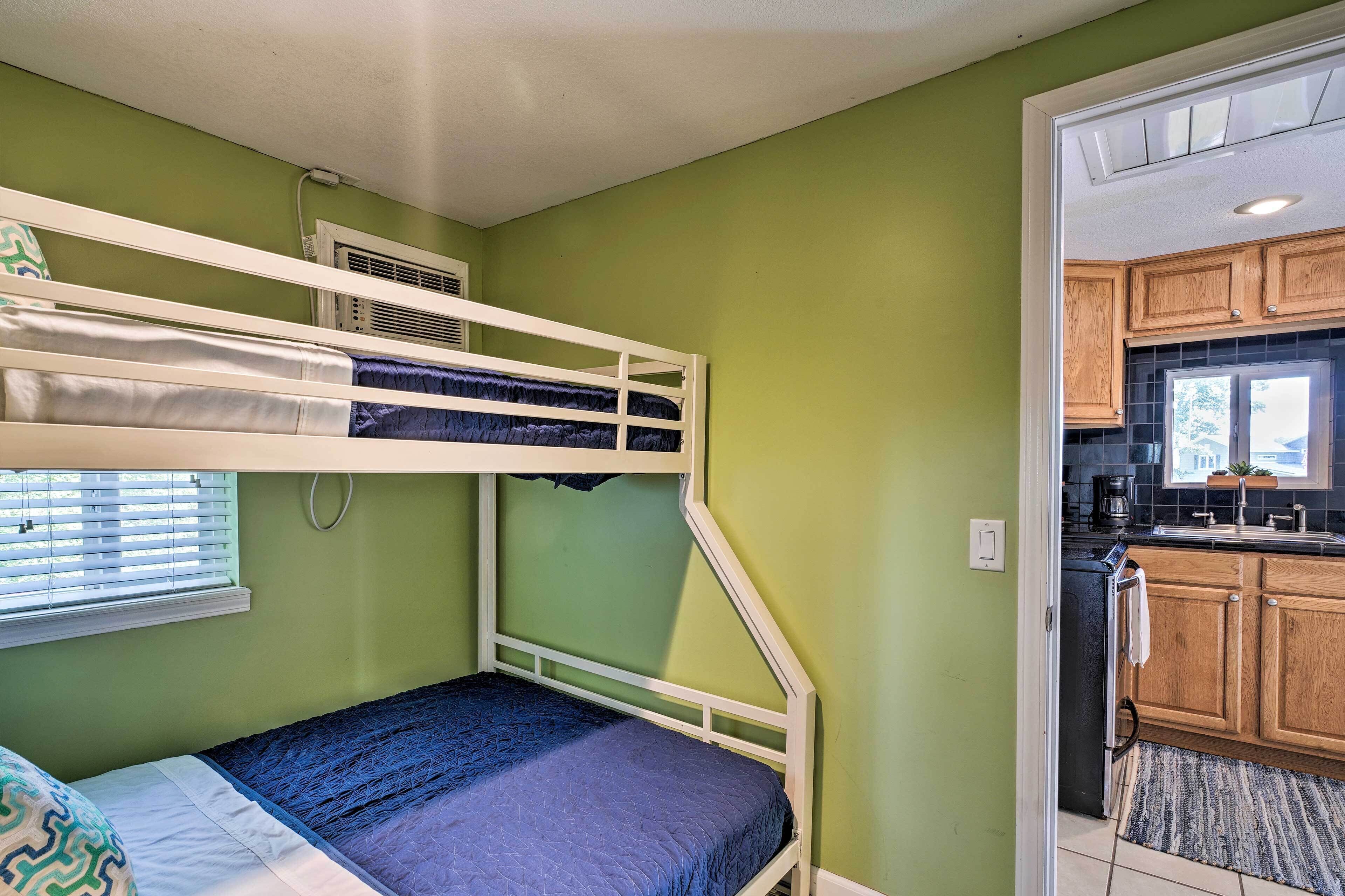 Un lit superposé avec un matelas bleu, une fenêtre avec des stores et un coin cuisine doté de meubles en bois et d’un évier.