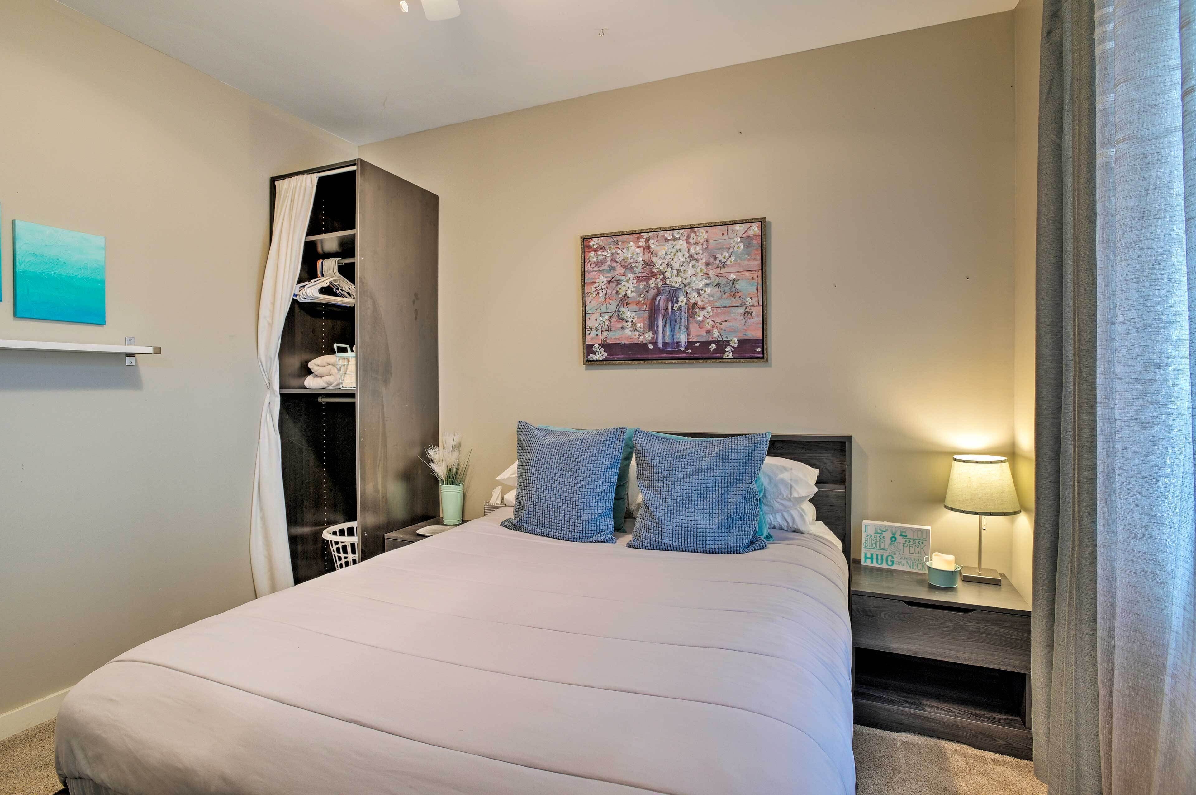 A neatly made bed with pillows, a bedside table with a lamp, a wall-mounted painting, and a closet with hanging clothes.