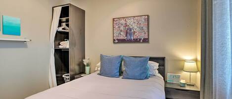 A neatly made bed with pillows, a bedside table with a lamp, a wall-mounted painting, and a closet with hanging clothes.