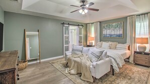 A bedroom with a bed, a chair, a nightstand, a ceiling fan, a window with curtains, and a painting on the wall. - Cozy-modern Pensacola Home: Large Yard, Grill (Pensacola)