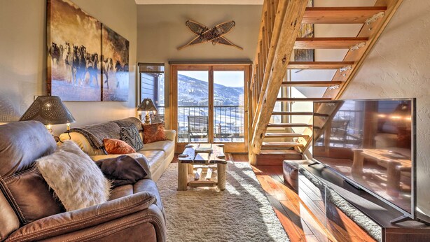 Un salon chaleureux avec un canapé en cuir, une table basse et un grand téléviseur. On peut apercevoir un escalier en bois et un balcon offrant une vue sur la montagne.