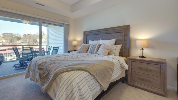 A bedroom with a large bed, a nightstand with a lamp, a window with blinds, and a view of the outside. - Water Views + Pool Access: Lake Ozark Escape! (Lake Ozark)