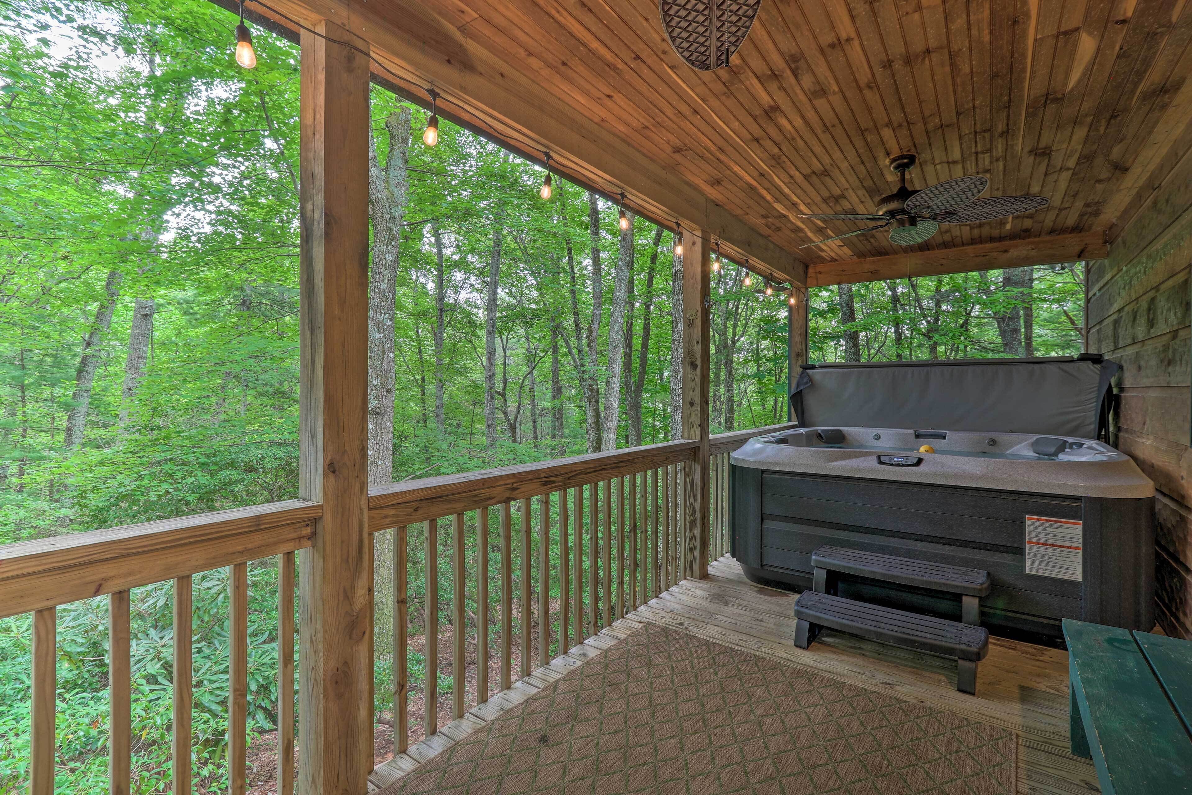 A wooden deck with a hot tub, a ceiling fan, and a view of a forest.