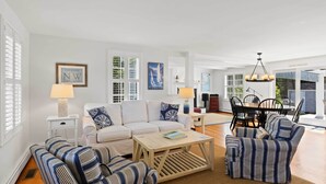 A spacious living room with a white sofa, striped armchairs, and a wooden coffee table. There is a dining area with a table and chairs, and a framed picture of a sailboat on the wall. - 3 Bicycles Provided! Cape Cod Cottage w/ Deck (Orleans)