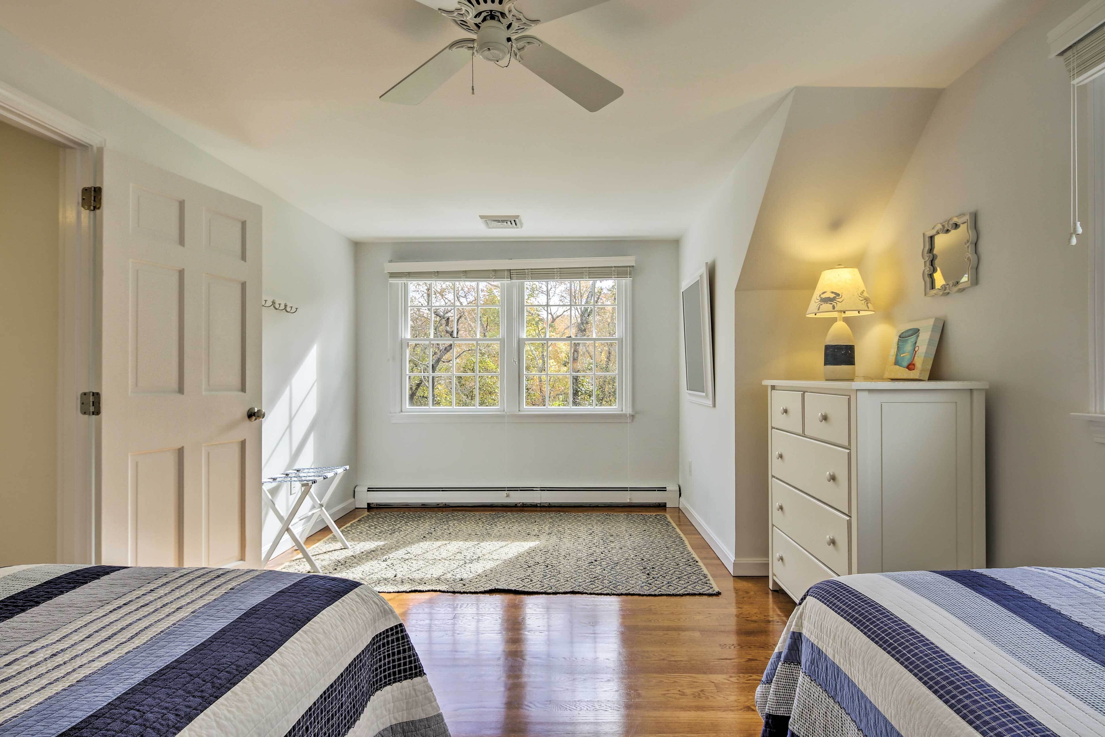 A bedroom with two beds, a ceiling fan, a window with blinds, a radiator, a dresser, and a lamp.