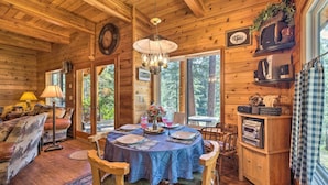A cozy cabin interior with a dining table set for four, surrounded by wooden furniture, a chandelier, and a large window offering a view of the forest. - Secluded Leavenworth Cabin on Chiwawa River! (Leavenworth)