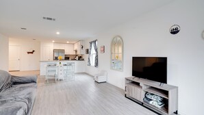 A modern living room with a grey sofa, a TV on a wooden stand, and a kitchen in the background. - Cozy Texas Escape Near Seaworld San Antonio! (San Antonio)