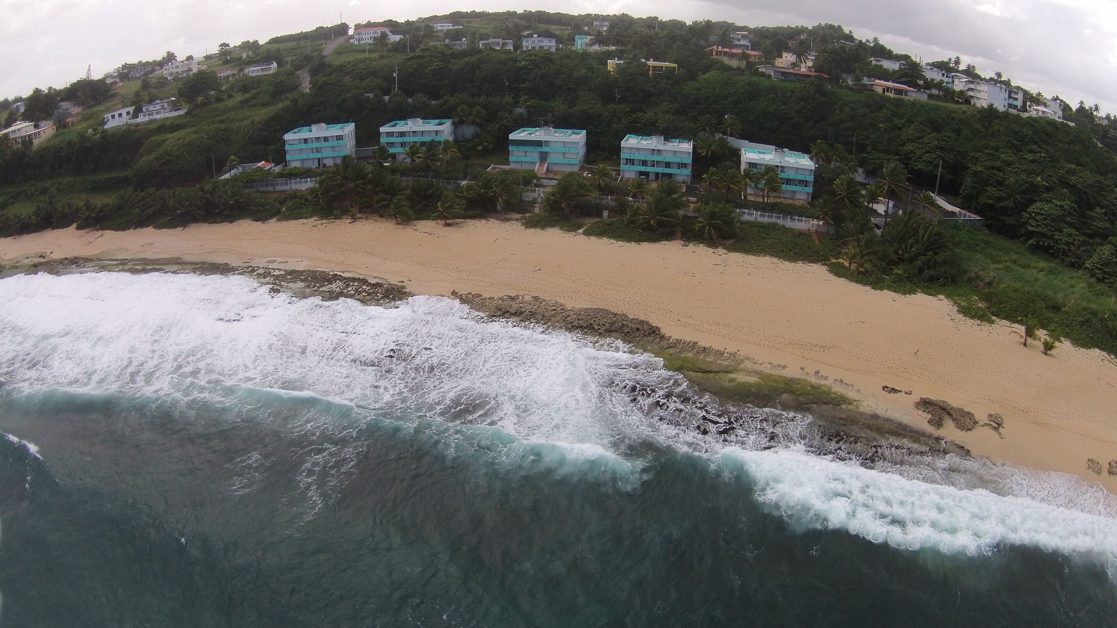 Vista costera con una playa arenosa y olas rompiendo en la orilla. En el fondo, varios edificios presentan un estilo arquitectónico uniforme, probablemente viviendas u hoteles.