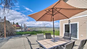An outdoor patio with a table and chairs, a large umbrella, and a view of a house and mountains in the background. - Ellensburg Home w/ Mountain Views on 3 Acres! (Ellensburg)