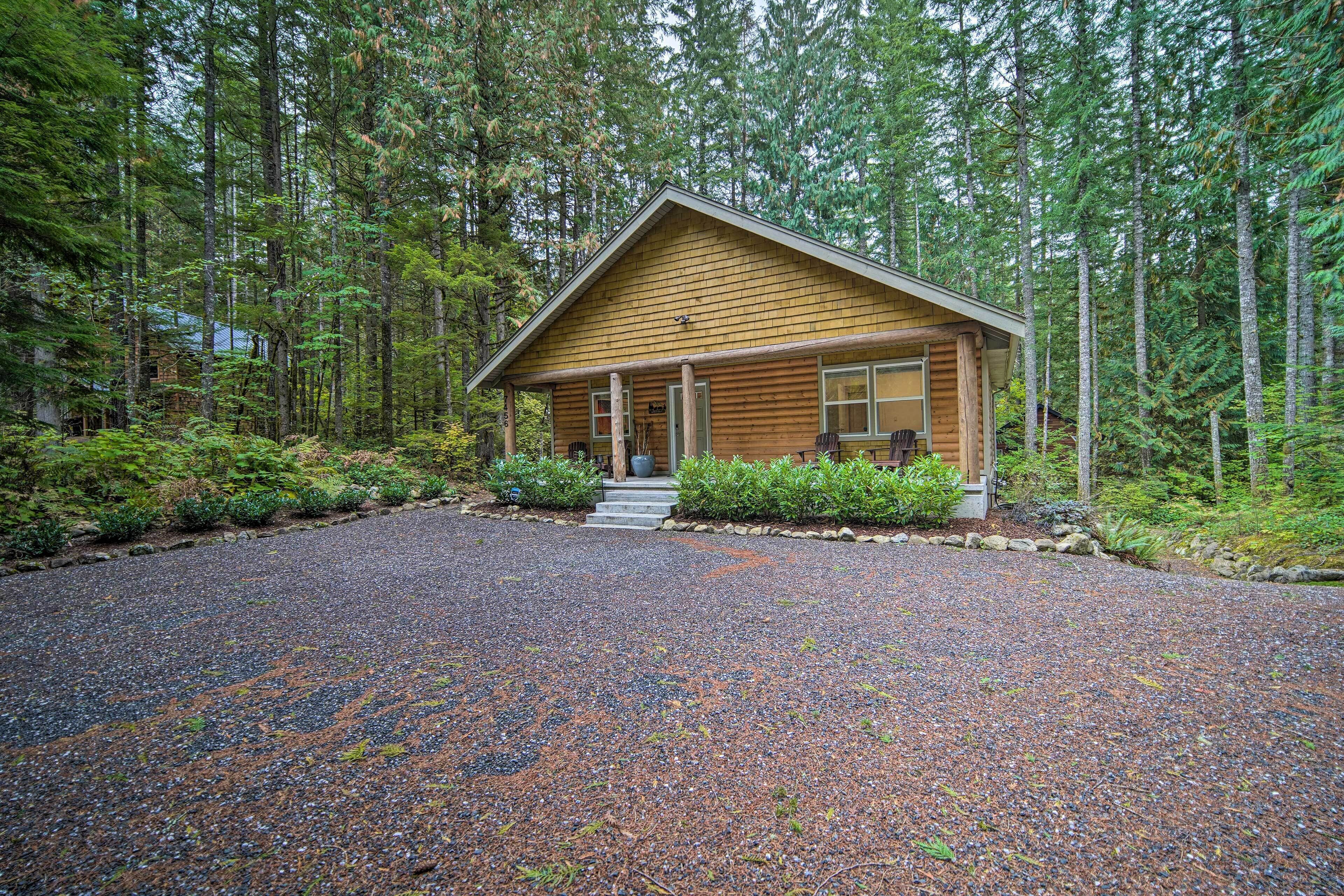A log cabin with a gravel driveway and a wooden porch, surrounded by dense forest.