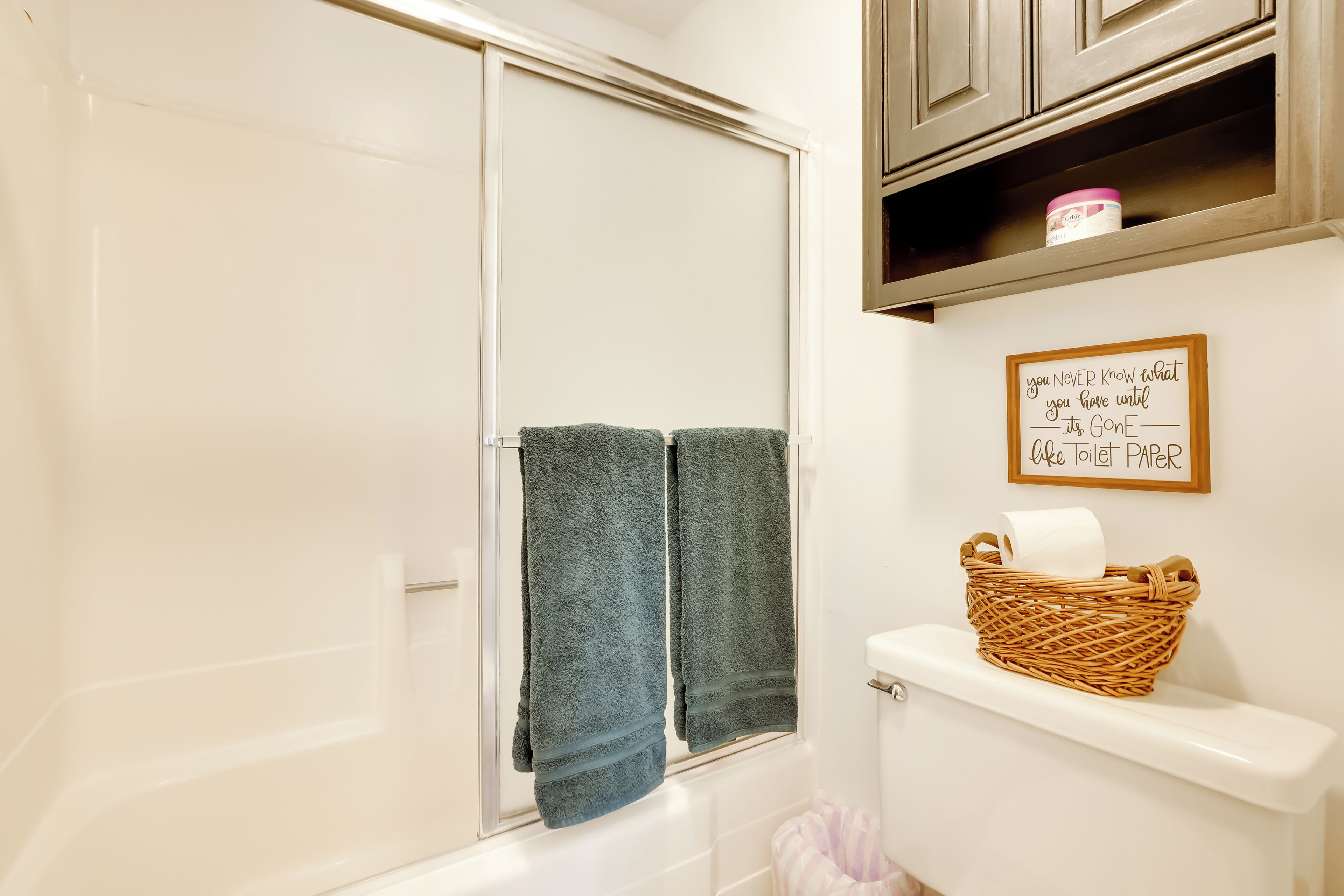 A bathroom with a bathtub, a toilet, and a towel rack with two towels. There is a basket with toilet paper on the toilet tank and a sign on the wall.