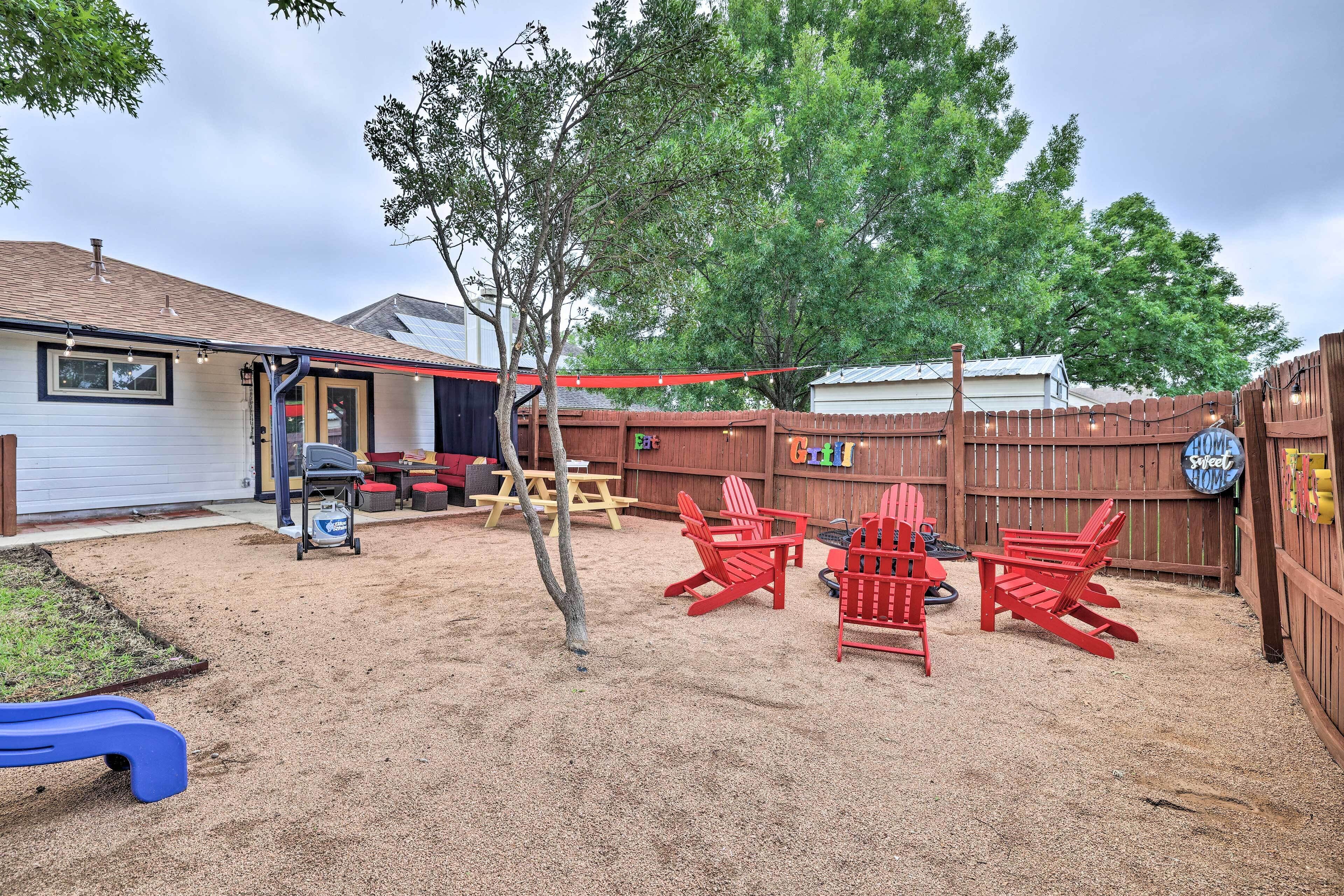 A backyard with a sandy play area, red Adirondack chairs, a wooden fence, and a house with a garage.