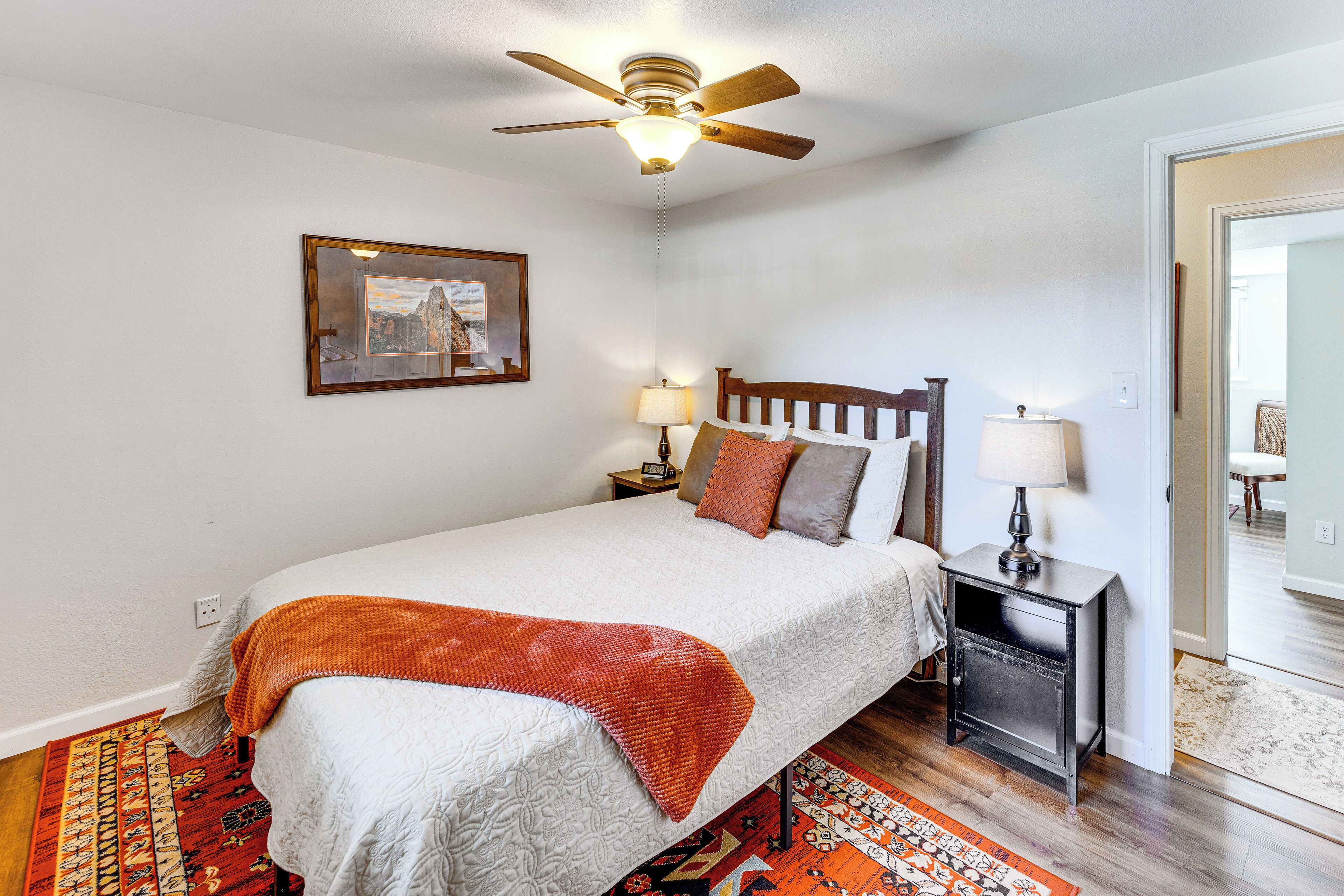 Une chambre à coucher avec un lit, un ventilateur de plafond, une table de chevet avec des lampes, un tableau encadré au mur et un tapis à motifs.