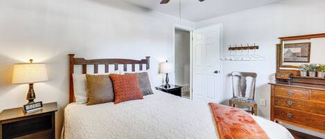 A bedroom with a bed, a nightstand with a lamp, a dresser, a ceiling fan, and a door.