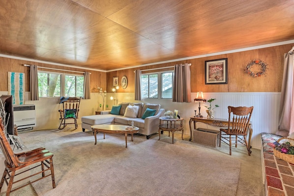A spacious living room with a sectional sofa, a coffee table, and a fireplace. There are two windows with curtains, a wooden ceiling, and a wall-mounted wreath.