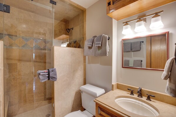 A bathroom with a glass-enclosed shower, a toilet, a sink with three faucets, and a mirror with lights above. - Bright Downtown Telluride Condo - Steps to Lift! (Telluride)