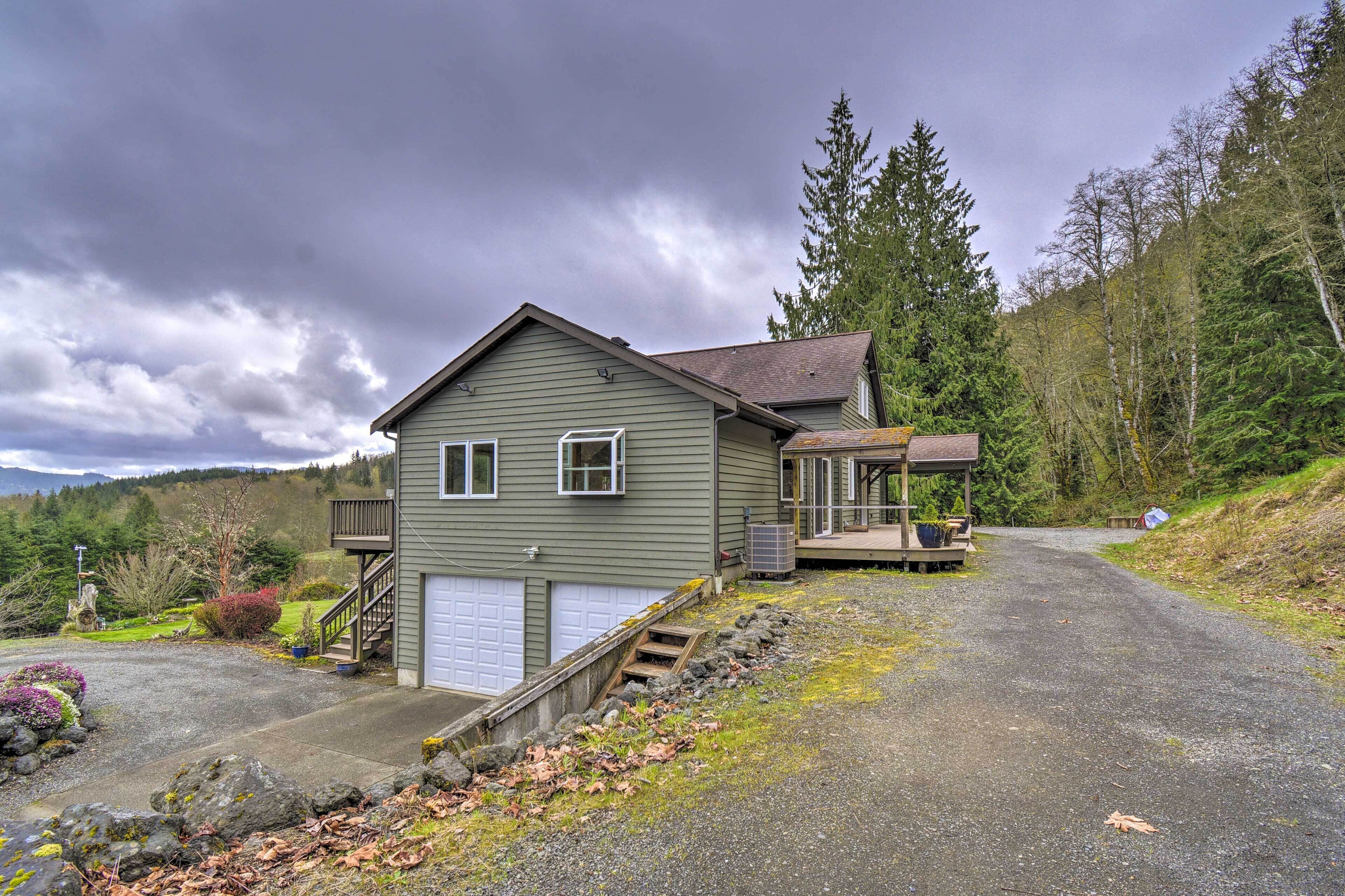 A two-story house with a garage, a covered porch, and a driveway leading to a forested area.
