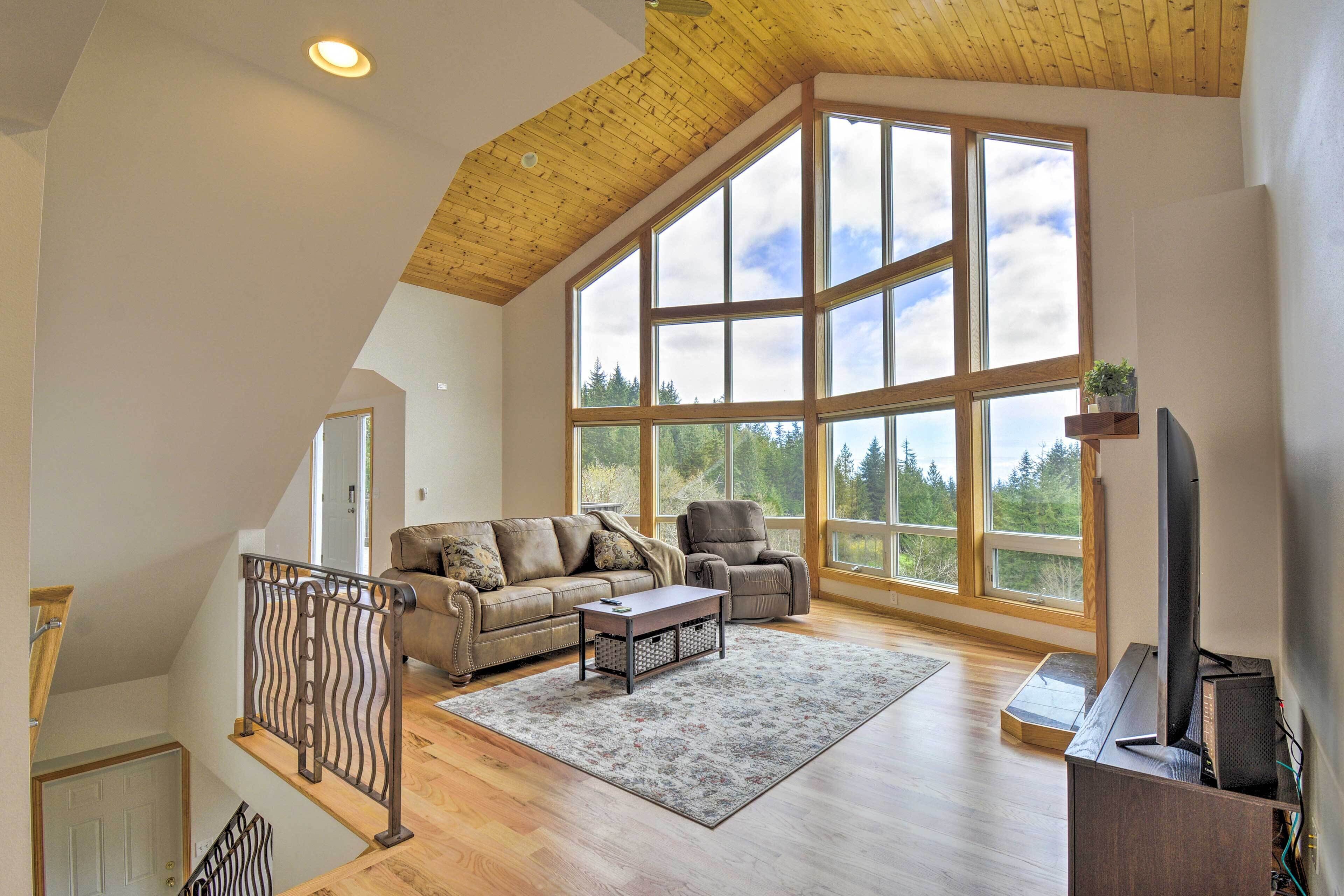 A spacious living room with a high ceiling, large windows, a wooden staircase, and a flat-screen TV.