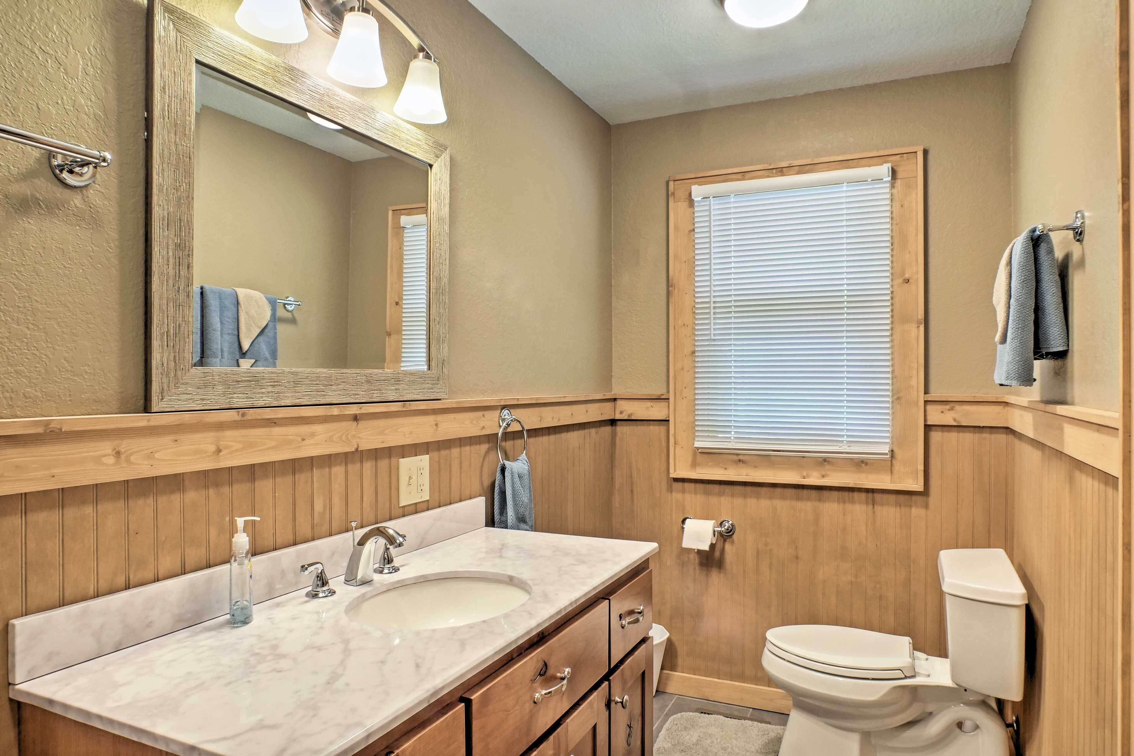 A bathroom with wooden paneling, a large mirror, a sink with a soap dispenser, a toilet, and a window with blinds.