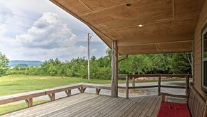 Een houten terras met een bank en een overdekt gedeelte, met uitzicht op een grasrijk landschap met bomen en een bergketen in de verte.