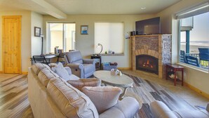 A living room with a stone fireplace, a large window with a view, a sofa, and a coffee table. - Quiet Waterfront Escape w/ Hot Tub & Beach Access! (Gold Beach)