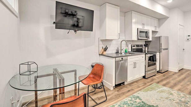 A modern kitchen with white cabinets, stainless steel appliances, a glass dining table, and a TV mounted on the wall. - Classy Philly Apartment: 2 Mi to Downtown! (Philadelphia)