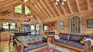 A wooden cabin interior with a kitchen, dining area, and living room furnished with leather sofas and a patterned rug. - Cabin w/ BBQ + Games - Walk to Blue Ridge Parkway! (Sylva)