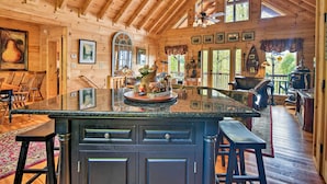 A wooden cabin interior with a large kitchen island, wooden ceiling beams, and a dining area with a bench. - Cabin w/ BBQ + Games - Walk to Blue Ridge Parkway! (Sylva)