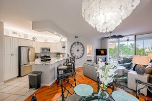 A modern kitchen with a central island, a dining area with round tables, and a living room with a sofa and TV.