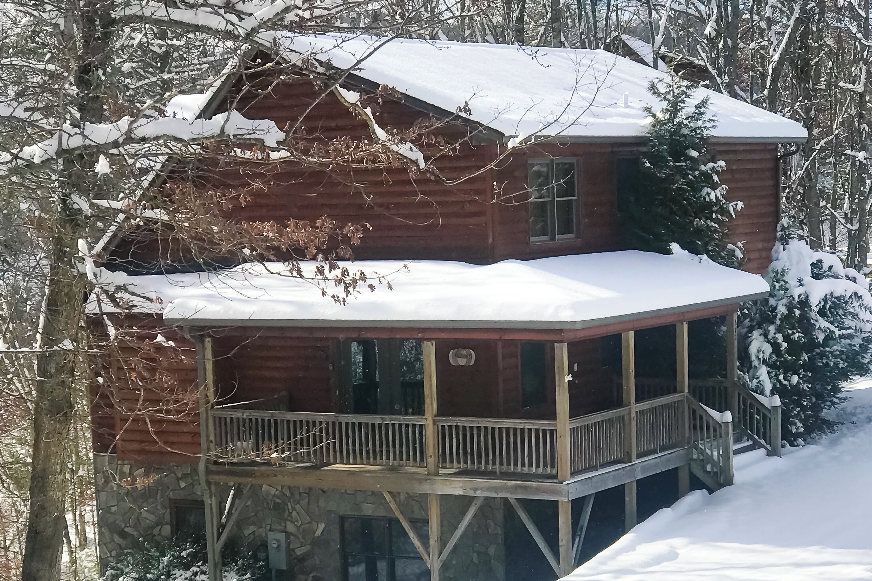 A wooden cabin with a covered porch and a stone foundation, surrounded by snow-covered trees.