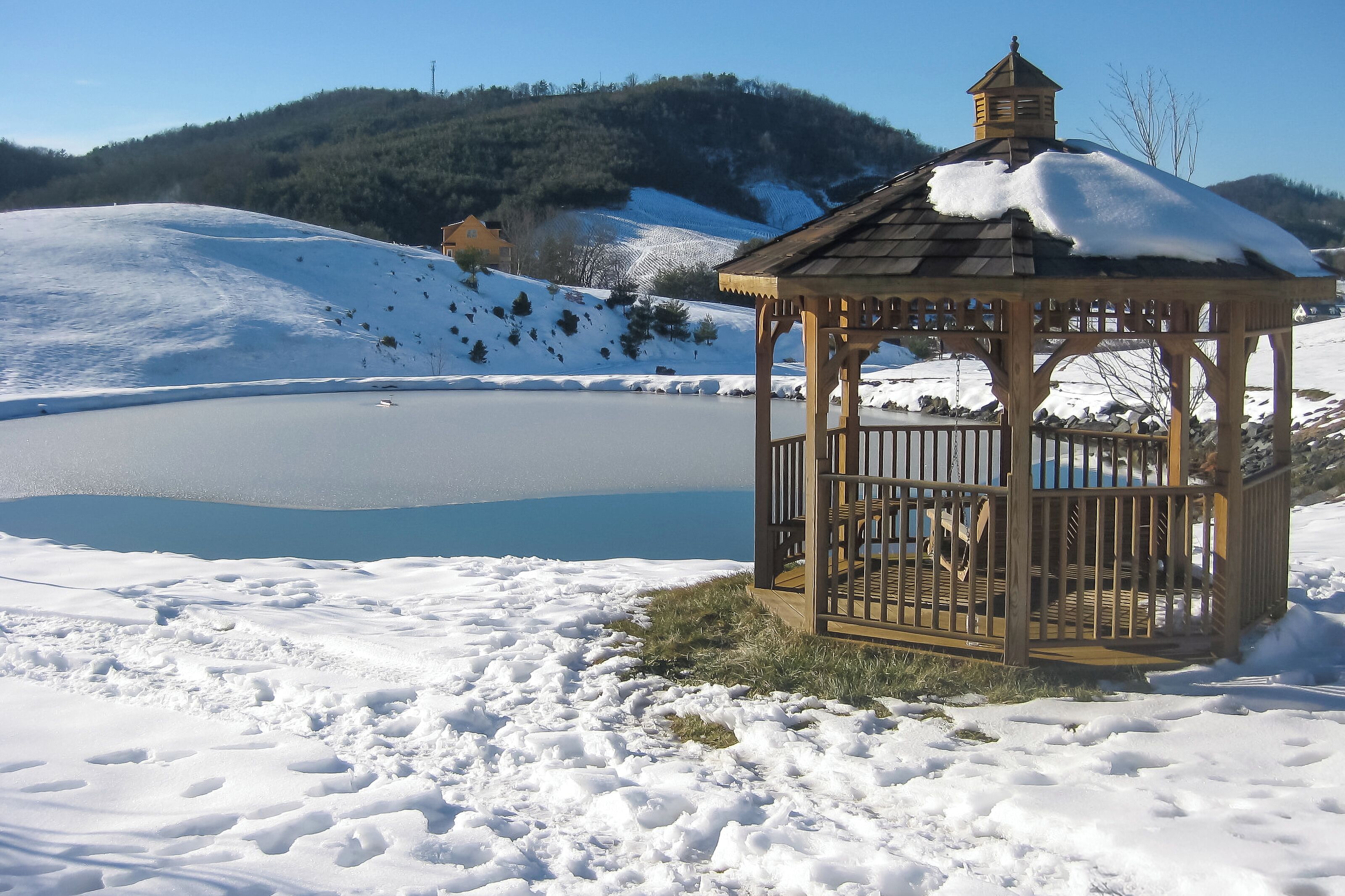 A wooden gazebo with a roof and railing, situated on a snow-covered ground near a body of water.