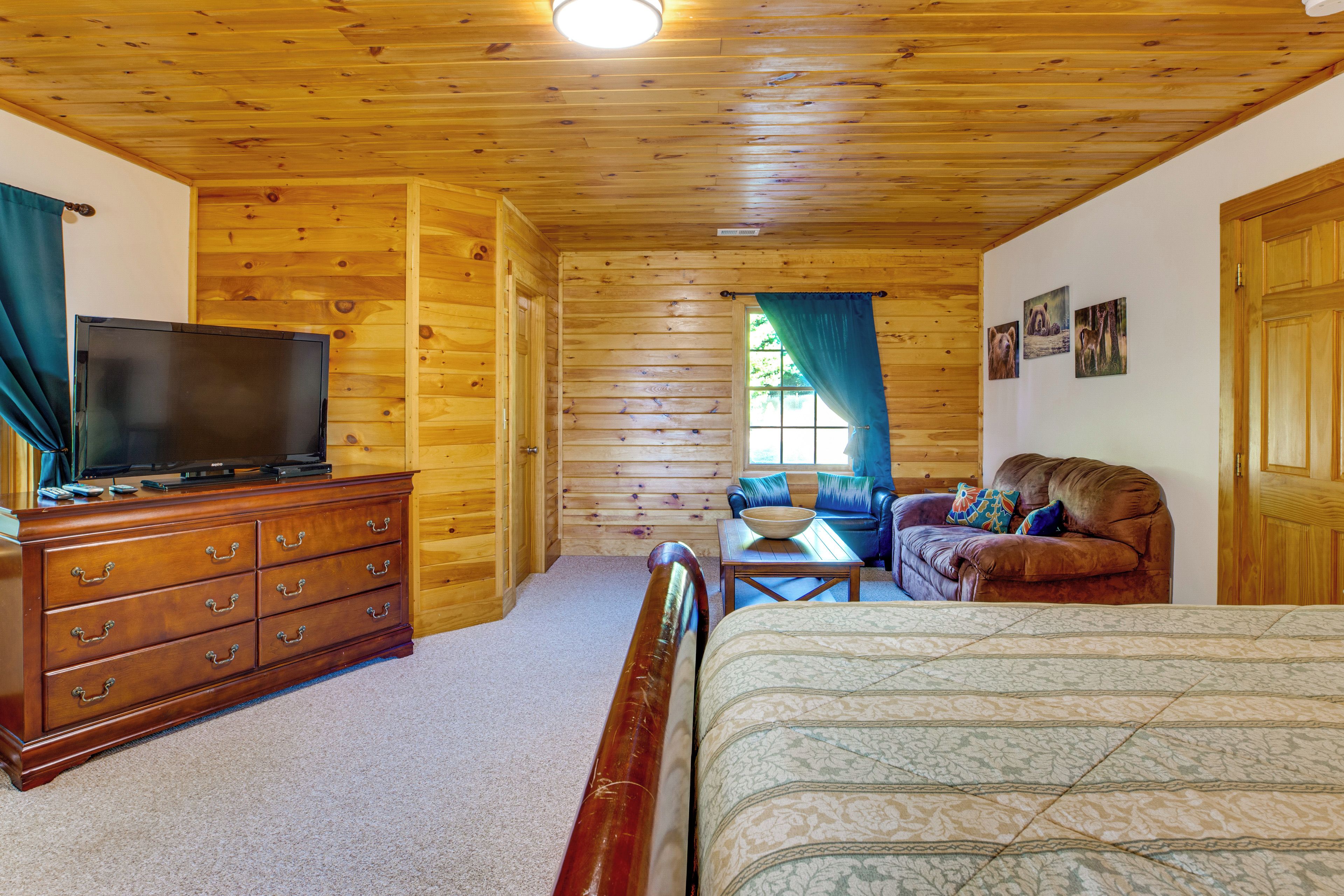 A log cabin interior with a wooden ceiling, a television on a dresser, a sofa, a bed, and a window with curtains.