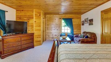 A log cabin interior with a wooden ceiling, a television on a dresser, a sofa, a bed, and a window with curtains.