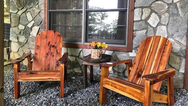 Twee houten Adirondack stoelen en een kleine tafel met een bloemenvaas, voor een stenen muur en een raam.