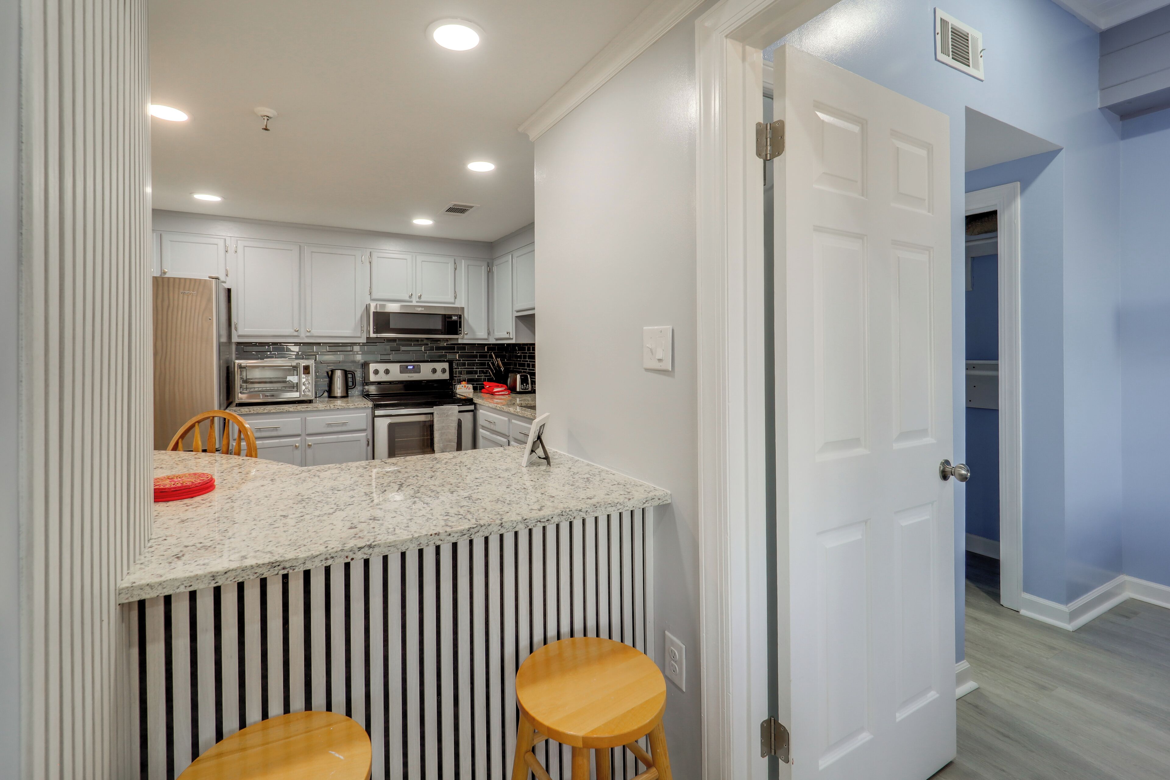 A modern kitchen with white cabinets, a granite countertop, and stainless steel appliances. There are two wooden stools and a red plate on the counter.