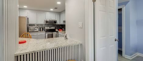 A modern kitchen with white cabinets, a granite countertop, and stainless steel appliances. There are two wooden stools and a red plate on the counter.