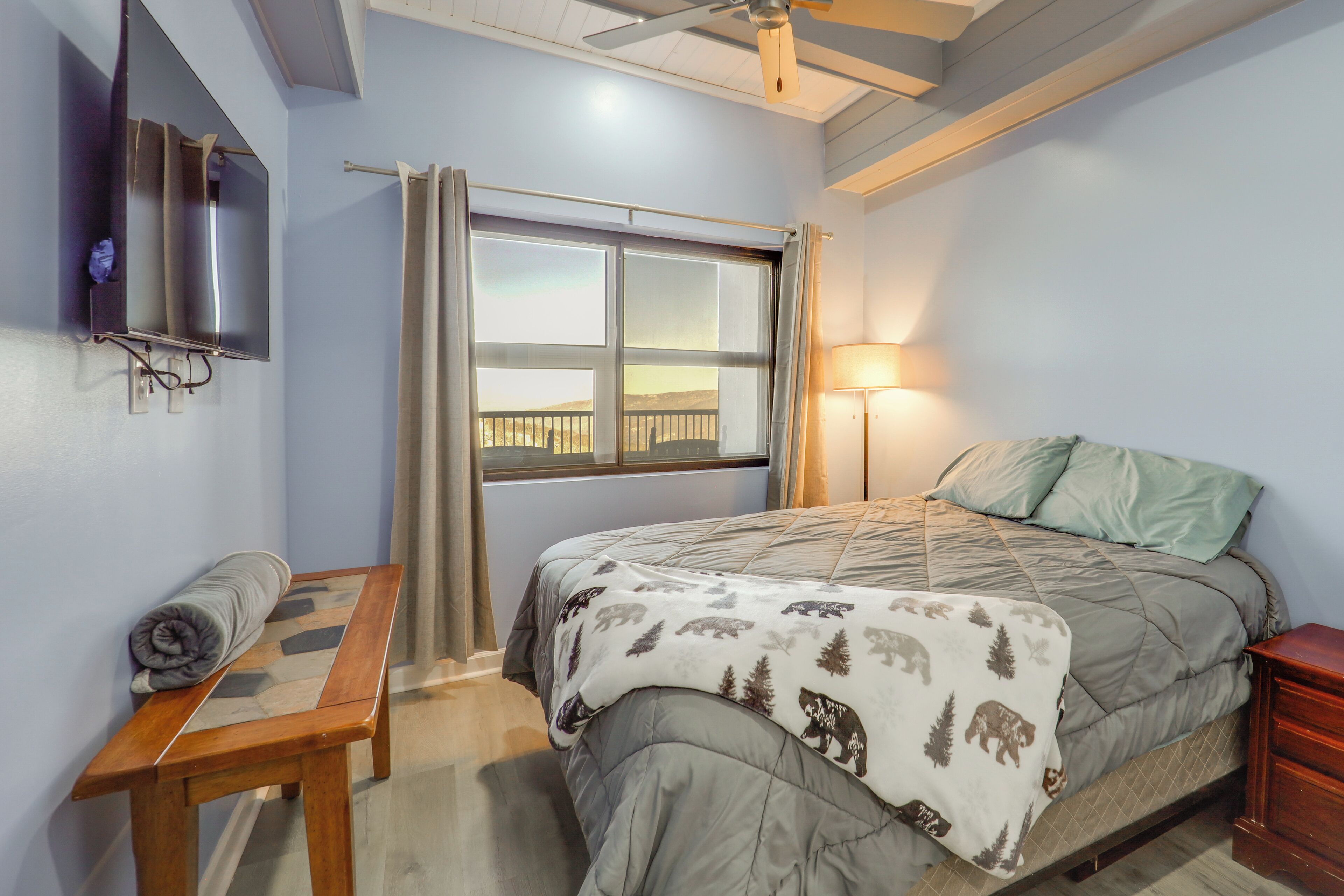 Un dormitorio con cama, televisor montado en la pared, ventana con cortinas y mesita de noche con una manta enrollada.