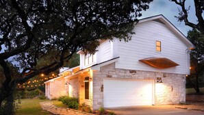 A two-story house with a stone foundation and white siding, featuring a garage and a covered porch with string lights. - Shaded Getaway w/ BBQ & Fire Pit in Austin! (Austin)