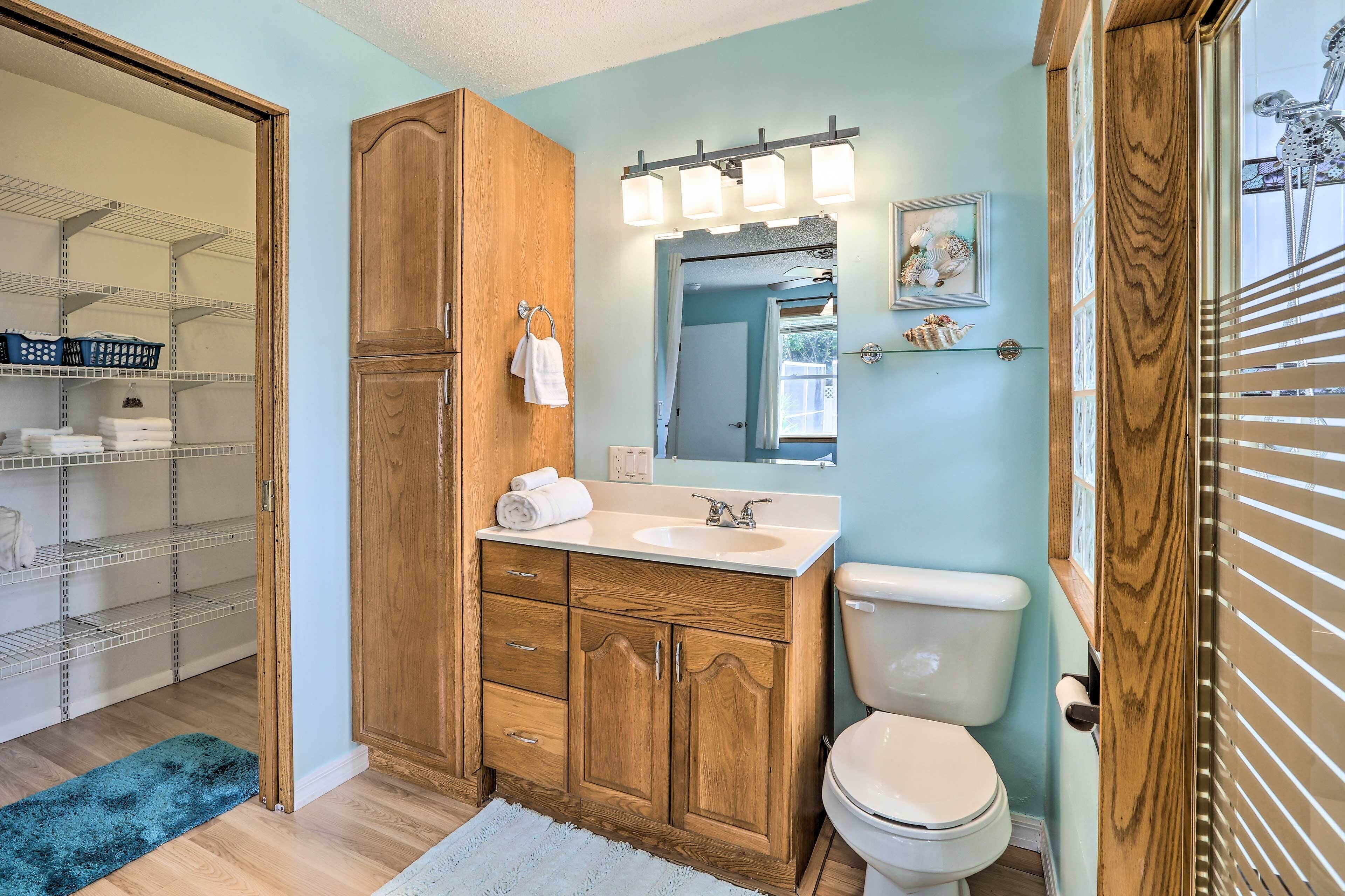 Ein Badezimmer mit Holzschränken, einem weißen Waschbecken und einer Toilette. Auf dem Boden liegt ein blauer Teppich und eine Ablage mit Körben und Handtüchern.